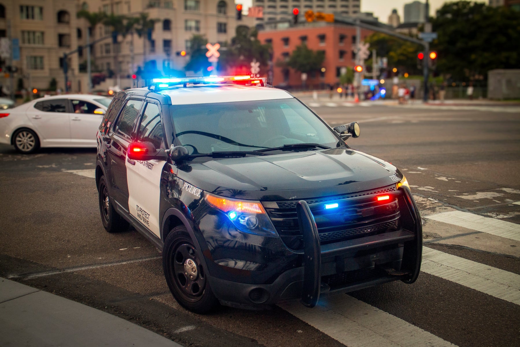 Police SUV with red and blue emergency lights activated, parked at an urban intersection in front of city buildings during evening traffic.